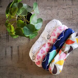 Cette photo représente un mélimélo de coussins relaxant pour les yeux ou eye pillow en coton garnis de riz et/ou lavande sur demande. Les imprimés vont du vert, au bleu en passant par le rose et le blanc avec des motifs divers. Une plante verte sert de décoration.