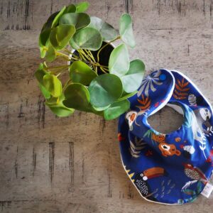 Cette photo représente un bavoir bandana vue de face en jersey fermé par deux boutons pression. L'imprimé est sur fond bleu roi lumineux avec des animaux de la jungle multicolores dans un dessin à l'esprit naïf. Une plante verte sert de décoration.
