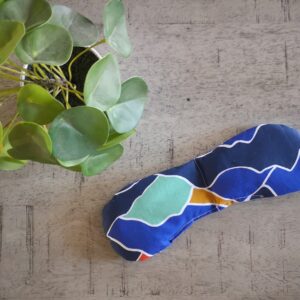 Cette photo représente un coussin relaxant pour les yeux ou eye pillow en coton garni de riz et/ou lavande sur demande. L'imprimé est sur fond bleu avec des motifs évoquant les montagnes aux couleurs vert eau, rouge, orange, bleu marine. Une plante verte sert de décoration.